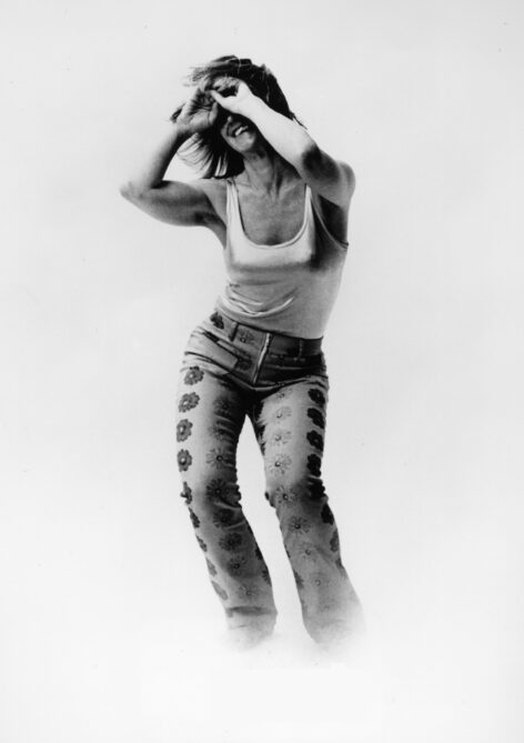 Iréne Hultman Monti standing with her arms covering her smiling face; she is wearing floral pants and a white tank top. Photo by Steve Ashmore.