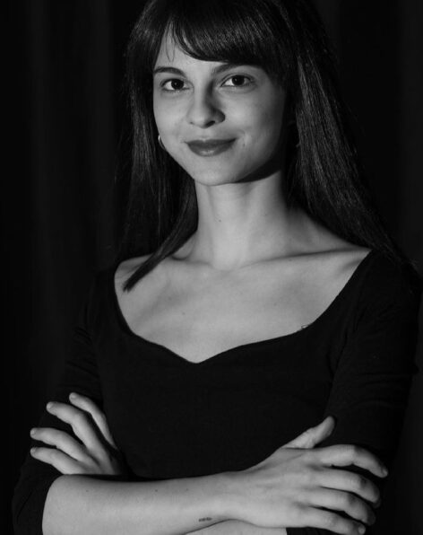A black-and-white portrait of a young woman standing against a dark background. She is wearing a black top with a wide neckline, her arms are crossed, and she has a calm, confident expression with a slight smile. Photo by Volkan Erkan