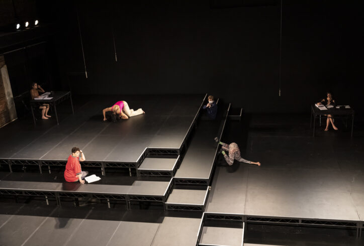 Remains performance photo; performers in various positions on a stage and risers, 2 sit at a table with papers in front of them. Photo by Rachel Papo.