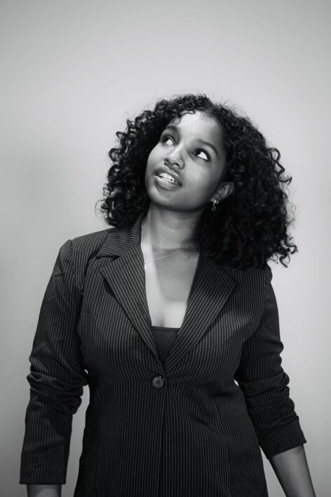 Black and white image of a dark-skinned woman with curly hair. She is wearing a pinstripe blazer, and looks up.