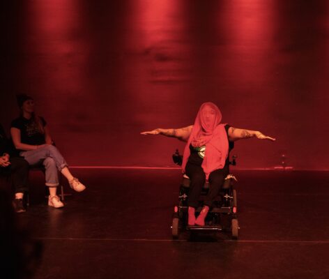 Under red lights, against a vermillion wall, with a crimson veil covering their head and face, NEVE is captured onstage, mid-dance, atop their power wheelchair. Their brown arms, peppered with tattoos, are outstretched to their sides, fingers extending, wrists slightly bent. Beneath the slightly transparent veil, there is a hint of large, kohl-lined eyes, a shapely nose and mouth, and black third eye antenna tattoos. At the edges of the photo, there is an audience, viewing the performance in the round. Photo by Michelle Smith-Lewis