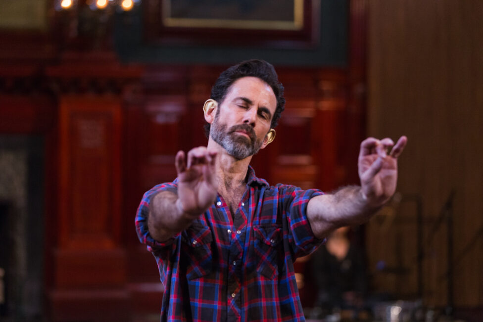 A male dancer with dark hair, closing his eyes as he raises both his arm with intricate finger position in an old building with red doors. The Set Up: Heni Winahyuningsih: PRINCESS by Wally Cardona, 2014 @ Park Avenue Armory, NYC
