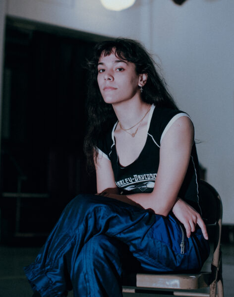 A pale woman with long dark hair and bangs sits cross legged with her arms folded. She is leaning slightly forward and has a subtle smirk on her face. She is wearing baggy blue ski pants and a tight black shirt. Photo by Olga Rabetskaya.