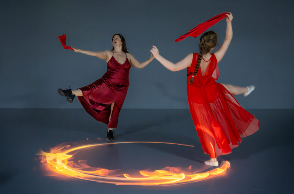 Two women stand in opposition to one another around a circle of fire. They both wear long red dresses and hold red scarves. One leg and one arm is out. Photo credit: Todd Carroll