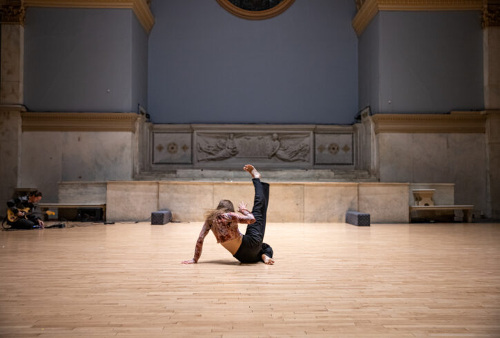 Sabina Bočková performing for Movement Research at the Judson Church. She sits with one leg extended upwards. Photo by Rachel Keane.