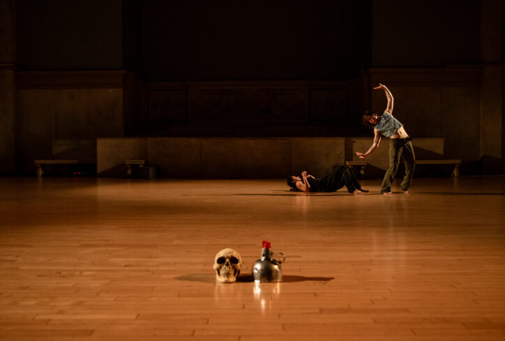 Photo from Punto de Inflexión/ Stephanie García and Jose Kolbeck. performing for Movement Research at the Judson Church. A skull and a red candle in a silver stand are a the foreground. In the background one dancer is on the floor and the other stands leaning sideways and spreading their arms wide.Photos by Rachel Keane