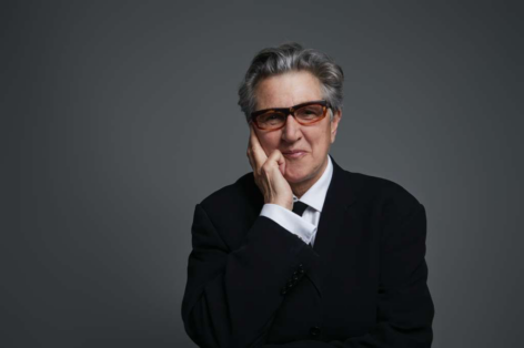 Peggy, a white woman with short hair and framed glasses smiles with one hand on their cheek; they are wearing a black blazer with a black tie and white shirt. Photo by Christa Holly.