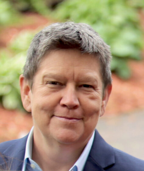 Eleanor, a white person with short hair smiles at the camera, they are wearing a blue blazer, behind them is blurred plant life. Photo by BFRESH Productions.