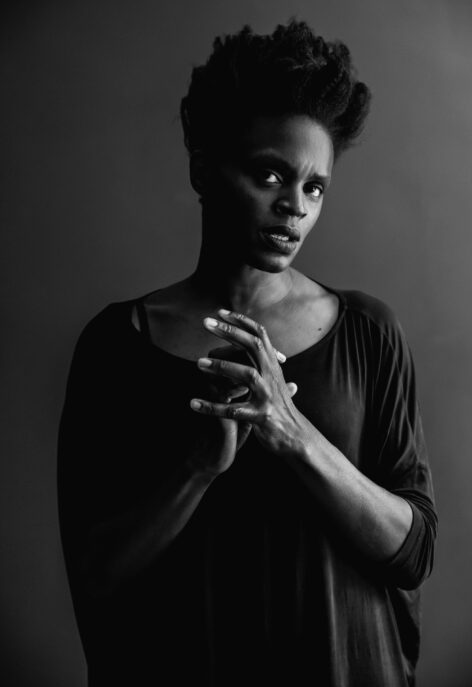 A black and grey photo of a Nigerian woman, Okqui in a loose black top, she looks at the camera, hands touching with her mouth slightly open. Photo by Michael Avedon.