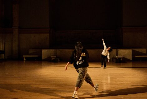A photo from Aleeza Garcia's piece for Movement Research at the Judson Church. Twi dancers move around the space one takes a wide step forward. The other slices their arm through the air. Photo by Ryutaro Mishima.