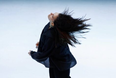 A woman wearing a black shirt is mid movement with her head tossed back against a white background. Photo by Ben McKeown.