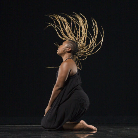 The image shows a dancer captured in a powerful moment on a dark stage. She kneels with her legs folded beneath her, leaning back as her head lifts upward. Her long, blonde braids sweep dramatically through the air, fanning outward in mid-motion. She wears a loose fitting black cami jumpsuit and is barefoot, her form highlighted by focused lighting against the black background. The contrast emphasizes the motion of her hair and the expressive shape of her body. The scene conveys strength, fluidity, and emotional intensity, evoking a sense of artistry and freedom in movement. Photo by Bill Frederking.