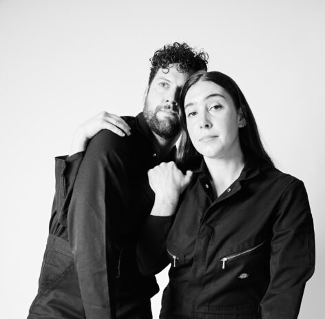 Black and white photo of Hollis and Nattie. They wear dark jumpsuits and embrace each other. Photo by Whitney Browne.