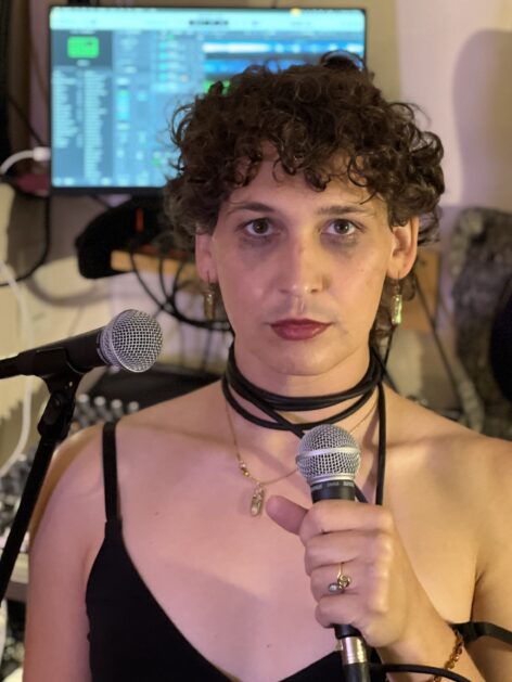 Margot stands looking into the camera in her music studio with overly dramatic mascara running down her face, dark red lipstick, and holding a microphone with the cable being worn as a necklace. Photo courtesy of the artist