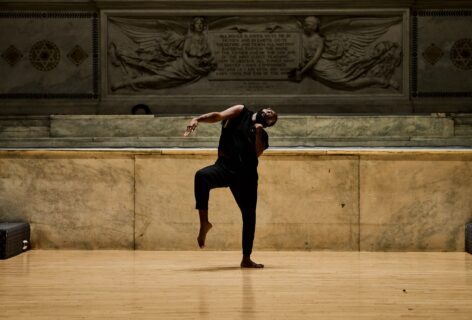 Jerron Herman performing for Movement Research at the Judson Church. Jerron stand son on leg balancing with their arm outstretched. Photo by Ryutaro Mishima.
