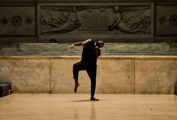Jerron Herman performing for Movement Research at the Judson Church. Jerron stand son on leg balancing with their arm outstretched. Photo by Ryutaro Mishima.