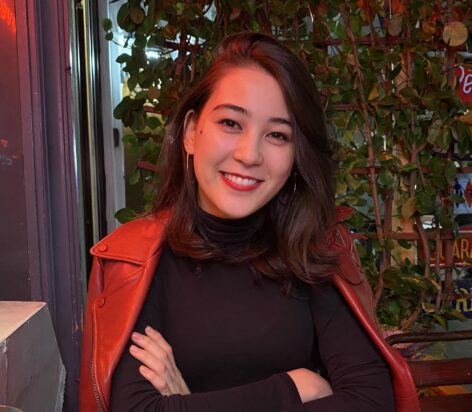 Smiling person with shoulder-length dark hair wearing a black top and a red jacket, sitting outdoors with greenery in the background. Photo courtesy of the artist.