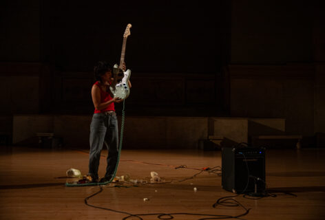 Photo from Yun Lee's performance for Movement Research at the Judson Church. Yun stands near an amp. There are wires and other objects on the floor. Yun stands wearing a red tank top and holding a blue electric guitar with the body of the guitar close to their head.