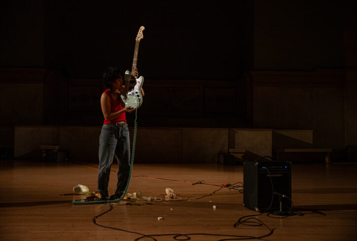 Photo from Yun Lee's performance for Movement Research at the Judson Church. Yun stands near an amp. There are wires and other objects on the floor. Yun stands wearing a red tank top and holding a blue electric guitar with the body of the guitar close to their head.