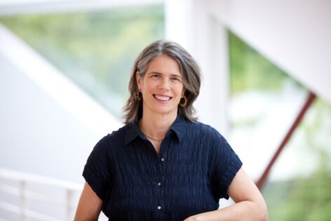 Katie, is a white woman with medium length hair in a dark blue top and brass ear rings, behind her is a blurred landscape. Photo by Argenis Apolinario.