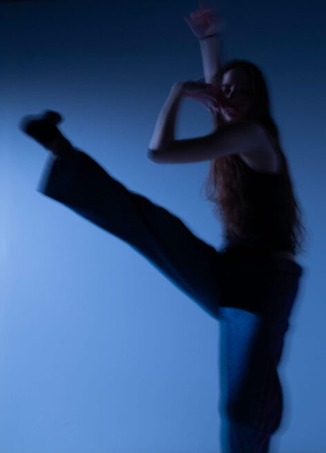 Dancer extends leg forward as arms contract into a boxy shape. The photo is tinted with a blue hue. Photography Credit: Sarah Kate Stolz.