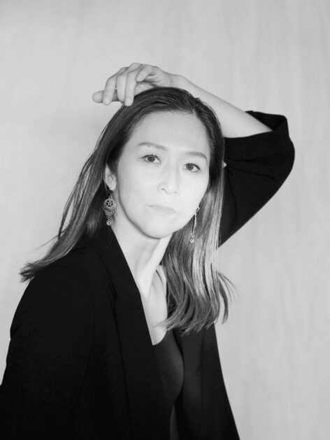 Monochrome portrait of a person with straight, shoulder-length hair wearing a dark blazer and top. They pose with one arm raised, hand resting on the head, creating a dramatic silhouette. Ornate dangling earrings catch the light. The plain, light background contrasts with the dark clothing, emphasizing the expressive posture and contemplative gaze. Photo by Hiroyasu Daido.
