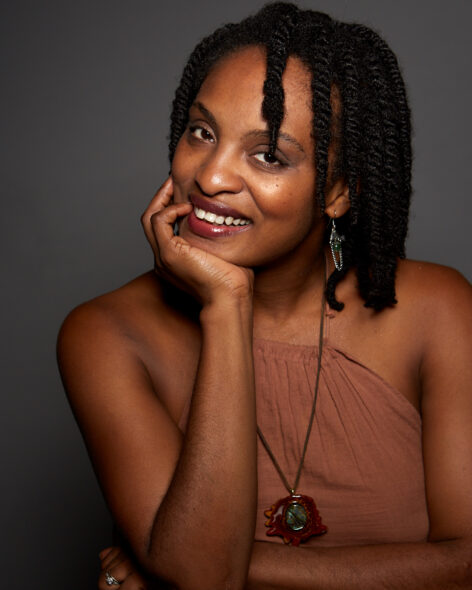 A close-up image of a woman with dark skin and her hair in short twists, wearing a brown strapless top and a necklace with a large, earthy pendant. She is smiling directly at the viewer with her right hand resting on her chin, against a solid gray background. She is also wearing earrings and a ring on her left hand.