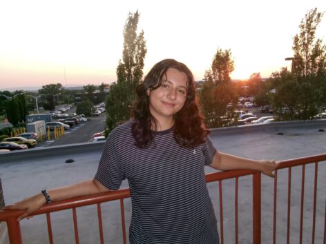 Portrait of Spencer Zambri-Moran. They are white, have long, dark brown, curly hair, and are leaning against a railing overlooking a sunset, wearing a navy and white striped tee shirt. Photography Credit: Bella Mantova.