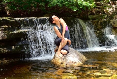 Water Spirit. A dancer perches on a large rock in golden-reflecting water. Their body seems to spiral as an extension of the rock, their face is turned upward towards the sunlight. A wide waterfall and bright green trees are behind them.Photo by Bea Ehrsam.