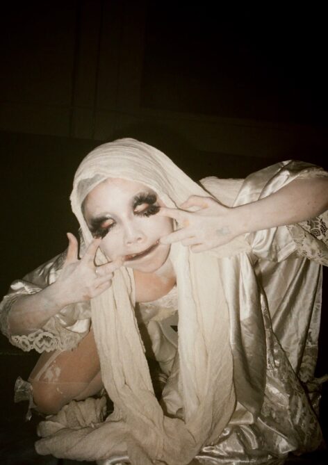 A portait of Yinx wearing white makeup and excessive eye makeup that assembles butterflies, pulling the sides of their eyes and mouth with their hands, in a white gauze costume. Photo/Image by Maoshiyi