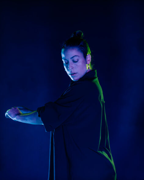 Zaquia, a woman with black hair pulled back, looks over her left shoulder towards the ground with arms extended in front of herself. She wears black clothes and her skin is colored by blue and green theater lights.Photo by Cam Kincheloe