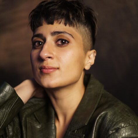 ankita, a light-brown skinned South-Asian femme with short dark hair looks directly at the camera with a slight smile. They are wearing a green, leathery, reptilian blazer, and their hand tucks gently behind their head in front of a brown background. Their eyes are slightly red. Photo by Bee Lively Photography.