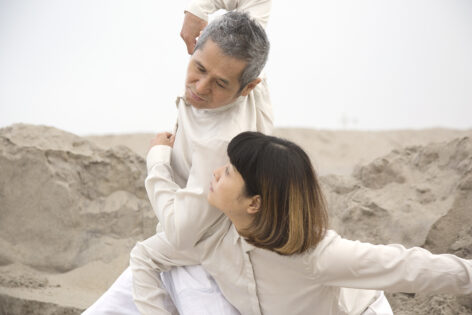 Two dancers perform a quiet duet on a sandy landscape. Dressed in light beige and white, they move with grace and connection—the woman supporting the man as they meet each other’s gaze. The soft tones of sand and sky create a calm, intimate atmosphere. Photo by Reggie Shiobara.