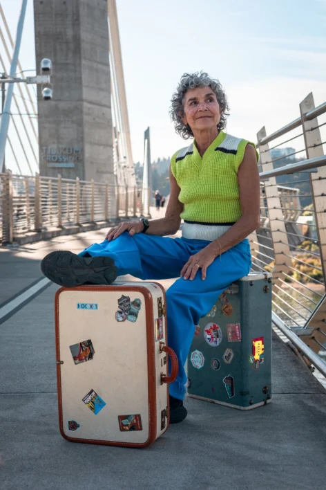 A dancer wearing bright blue pants and lime green vest performs with two vintage suitcases on a bridge in Portland, OR.  Photo by Ashton Bisner.