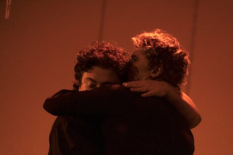 under red stage lighting, mik and tor, two white dancers embrace each other. Photo by James Izlar