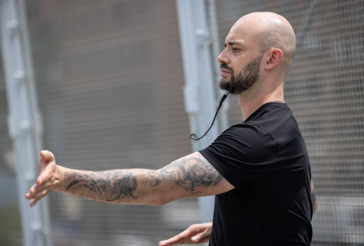 Christopher extends his right arm in the anterior-posterior plane. His long braided beard spirals around itself. His arms are heavily tattooed and he wears a black tee. Photo by Walter Wlodarczyk.