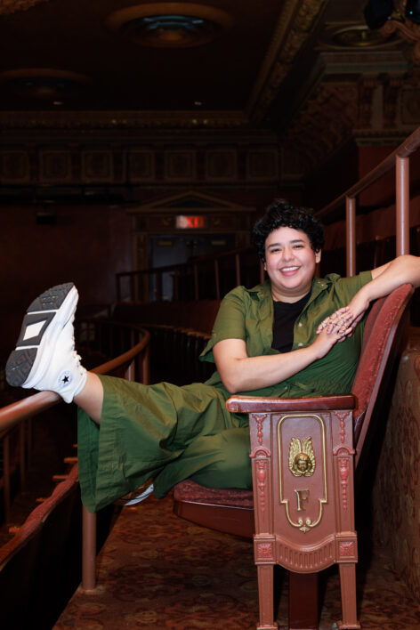 A photo of Maya Quetzali Gonzalez. She sits in a wooden theater seat wearing a green jumpsuit. She sticks a leg out in front of her and smiles brightly. Photo courtesy of the artist.