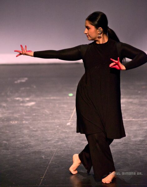 A photo of Sukanya wearing a black tunic and black flowy pants with left arm stretched out with a flower mudra/gesture hand (and right hand (painted red) at chest level with a bee gesture. The left leg is crossed in front of the right leg and the left foot is stretched off the floor. Photo Credit: DSJPhotography.