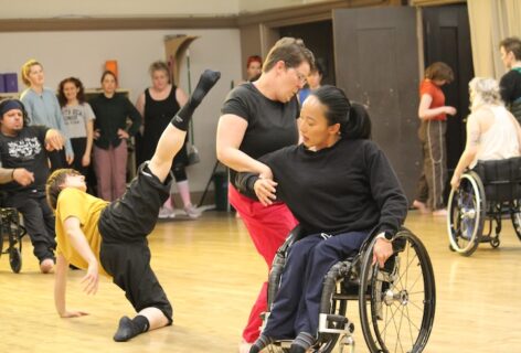 Three disabled and d/Deaf dancers improvise together in a well-lit studio space. A group of disabled, non-disabled, d/Deaf, and neurodivergent dancers observe their exploration in the background. Photo/Image by Nadia Adame.