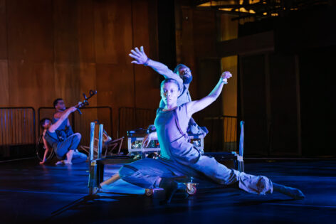 AXIS dancer, A(laj)a, lunges in front of a hexapod robot that company dancer, Tebandeke, is perched on top of. A(laj)a's left arm and leg are full extended to the side of their body while Tebandeke reaches and rotates his right arm over A(laj)a's head. There are two company dancers in the left of the background; one seated on the ground, one in a wheelchair, extending two crutch handles towards the upper right diagonal of the image.