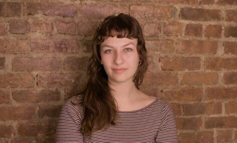 Photo of Alma's face in front of a brick wall. They have medium length brown hair with bangs, and wear a red and grey striped shirt. 