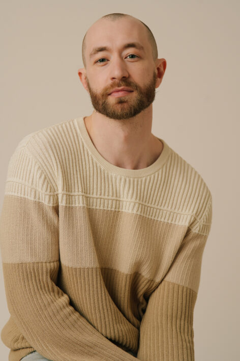 Alex Gibson posing for a portrait. They have a beard and short cropped hair. They wear a knitted sweater with three shades of beige in stripes. Photo courtesy of the artist.