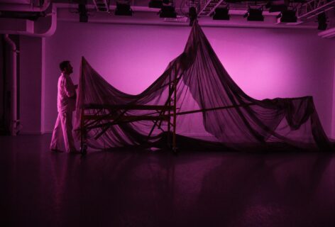 This image is taken in a dance studio. A person dressed in white is pulling a large object through the space. The space is bathed in pink light.