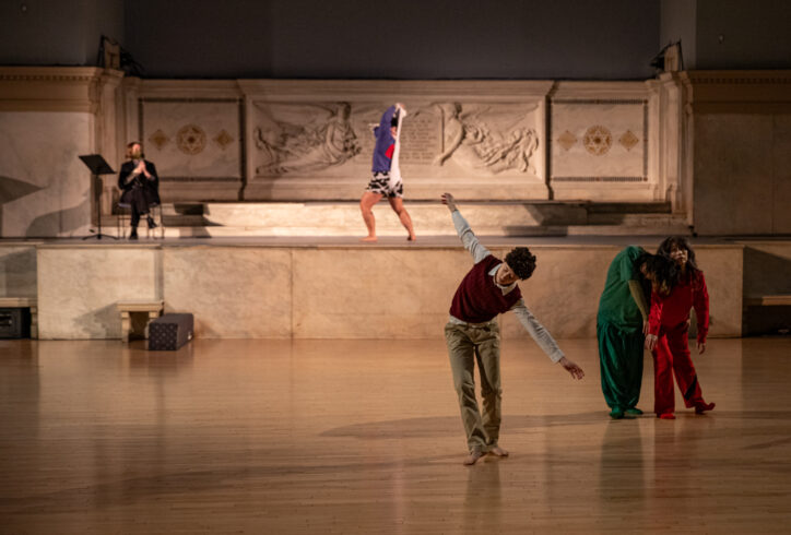 Lu Yim's work for Movement Research at the Judson Church. Dancers move around the space. One dancer in the forefront stands leaning sideway with their arms outstretched. Two dancers one in green and the other in red share weight. At the altar a person wears all black and sits near a music stand. Another person stands in a slight lunge with hands in their head pulling a white fabric Photo by Rachel Keane.