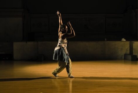 Photo of New Kyd performing for Movement Research at the Judson Church. They're caught mid-step and reaching upward with one arm while the other bends at the elbow and reaches in front of their face. Photo by Ryutaro Mishima.