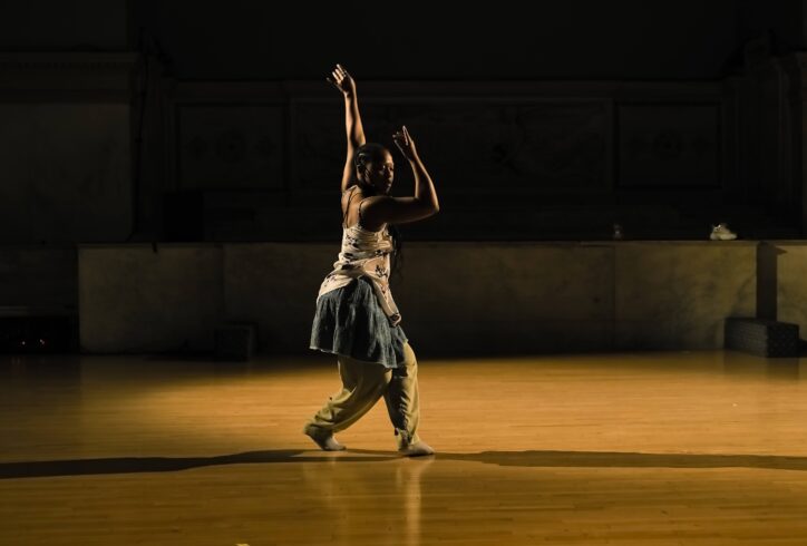 Photo of New Kyd performing for Movement Research at the Judson Church. They're caught mid-step and reaching upward with one arm while the other bends at the elbow and reaches in front of their face. Photo by Ryutaro Mishima.