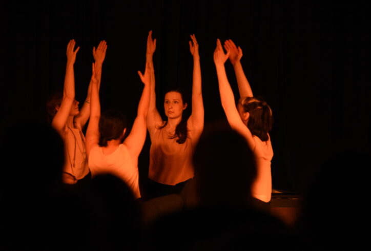 Four dancers with their arm extended up in a seated cross legged position with tan tank tops. Photo courtesy: DSJPhotography.