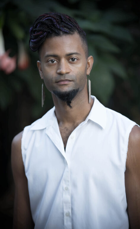 A photo of Ron Ragin. He wears a white button down sleeveless top. He has a short beard and wears long earrings. He looks into the camera with a neutral expression. Photo courtesy of the artist.