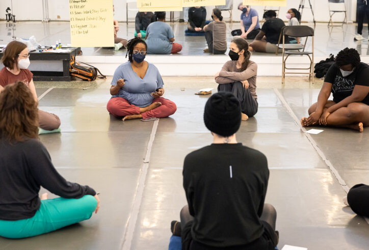 Kayla, a thick Black dancer with dark locs over the right side of their head and clear-framed glasses, sits in a circle with a group of seven dancers, some visible in the room, and some in the mirror. Everyone wears face masks and has their body oriented toward Kayla, as Kayla speaks animatedly with their hands, eyebrows raised. Alison, a white queer femme and access coordinator for Circle O – also an avid swimmer in the waters of Lake Michigan – wrote this description with a strong cup of breakfast tea next to her. Photo credit: Lynn Lane.