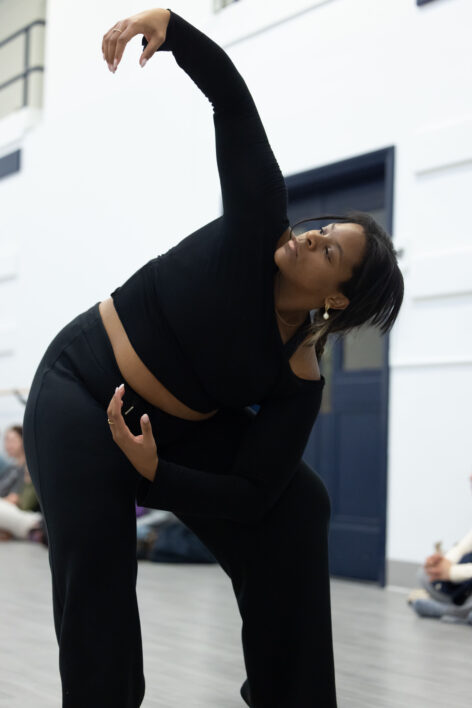 Katia is standing, leaning over her left knee and opening her torso towards the ceiling. She gazes upward at her uplifted right hand, as her other rests below her waist.